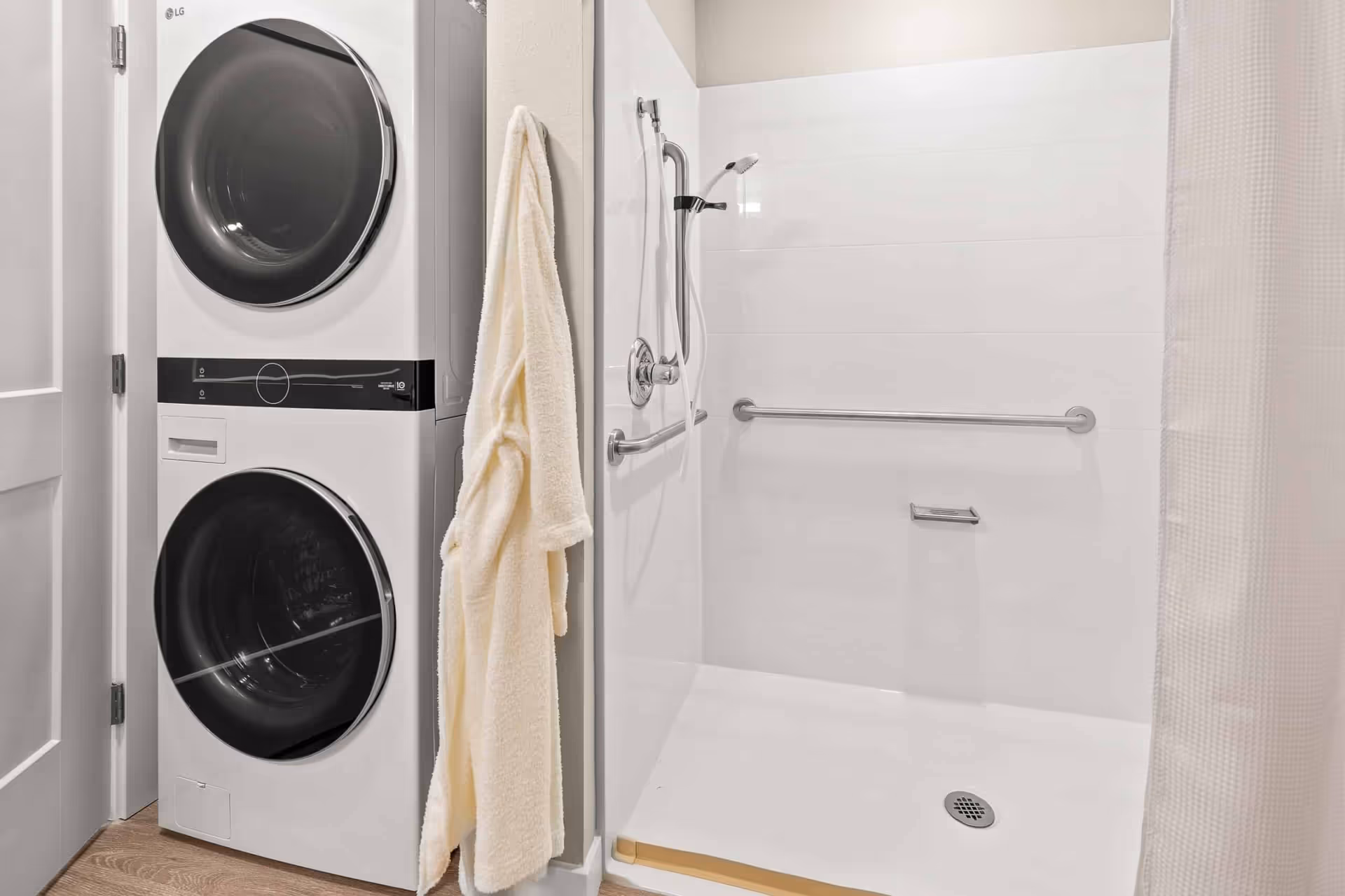 A modern bathroom featuring a walk-in shower with a handheld showerhead and grab bars, next to a stacked washer and dryer unit. A cream-colored bathrobe hangs on a hook between the shower and the laundry appliances.