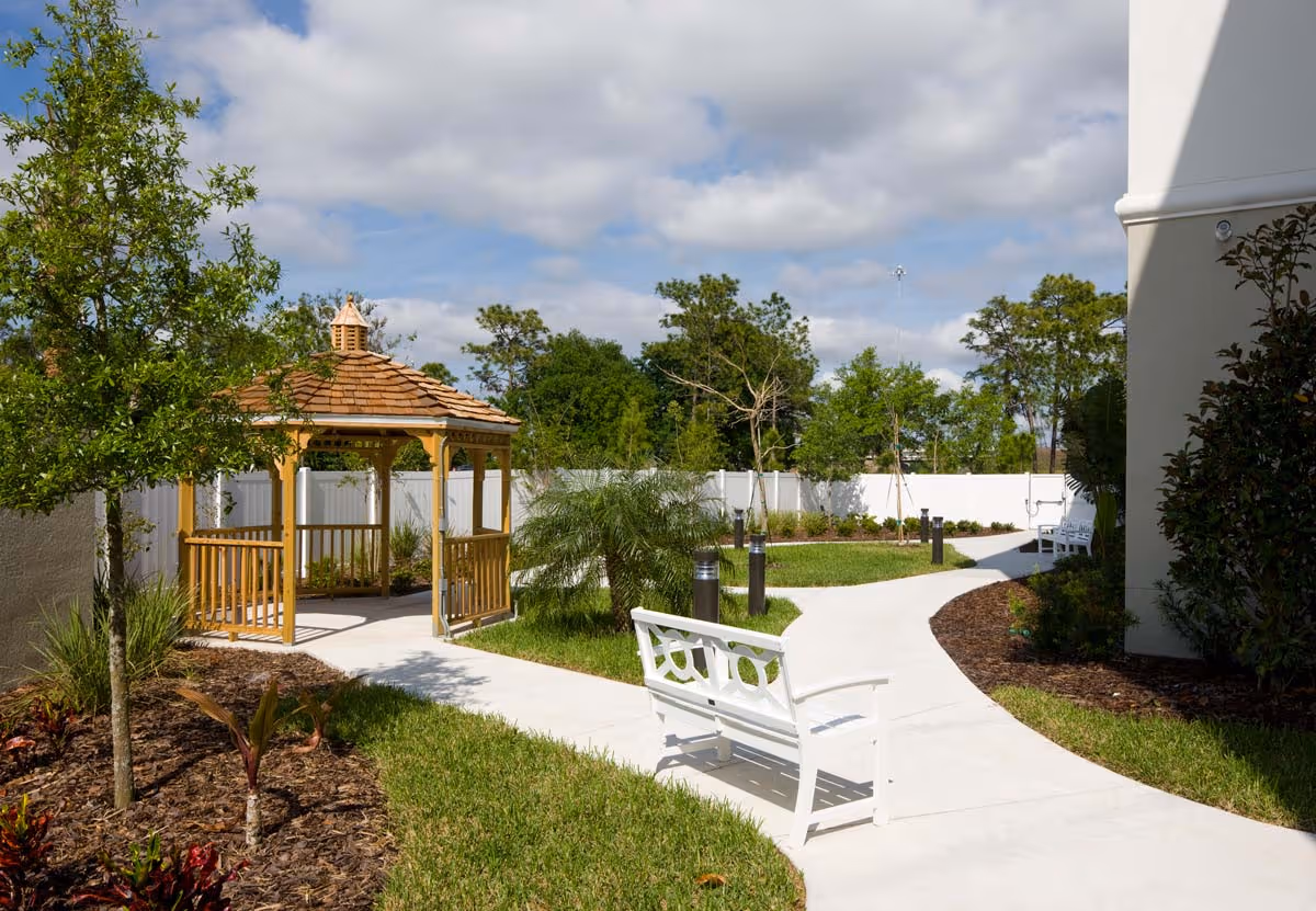 Outdoor courtyard with a wooden gazebo, white benches, and a winding concrete path surrounded by landscaping and trees.