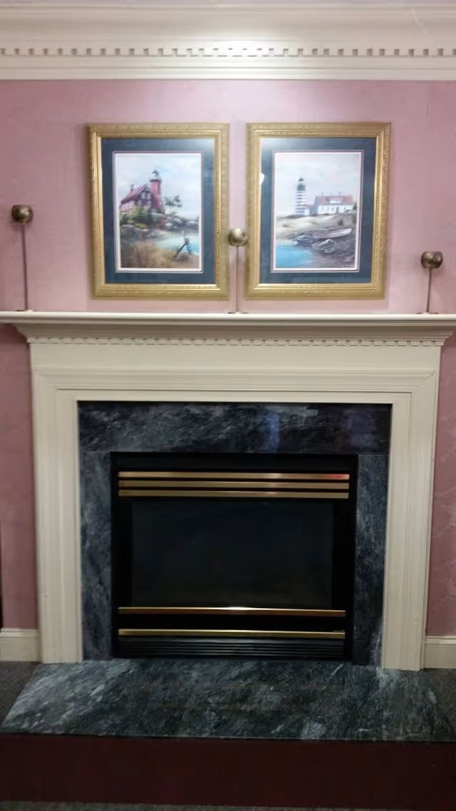 A decorative fireplace with a black and gold framed firebox surrounded by a marble hearth and white mantel. Above the mantel, two framed paintings of lighthouses hang on a pink wall, with two brass candle holders mounted on either side of the paintings.