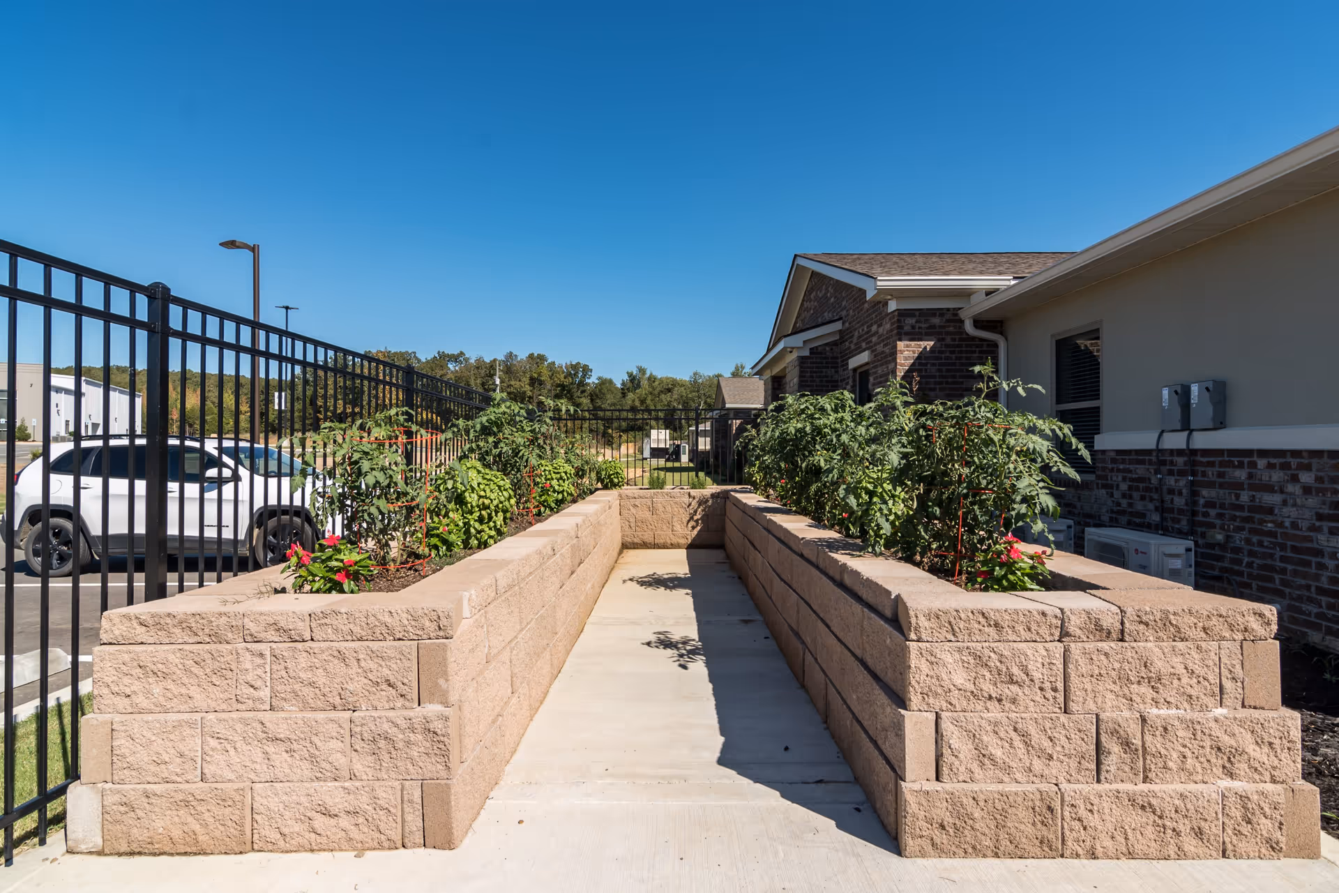 Outdoor garden area at Crestview Senior Living featuring two raised stone planter beds with green plants and flowers, a black metal fence, a white car parked nearby, and a brick building under a clear blue sky.