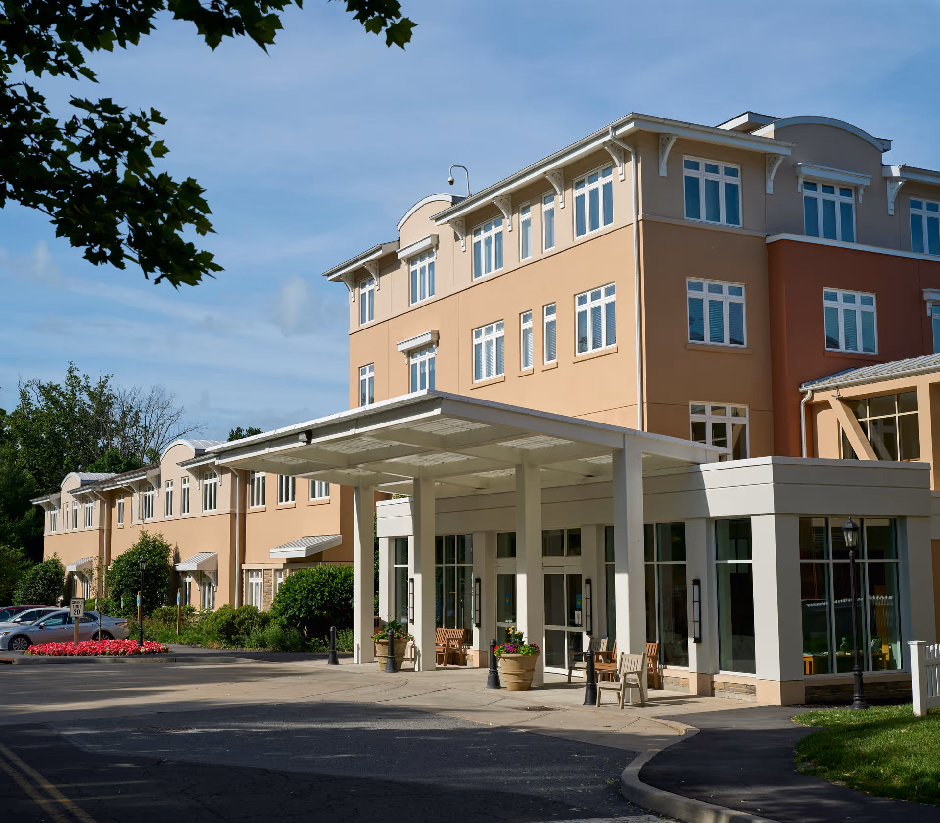 Exterior view of a multi-story senior living facility building with beige and light brown walls, large windows, and a covered entrance with benches and potted plants. There are trees and parked cars visible nearby under a clear blue sky.