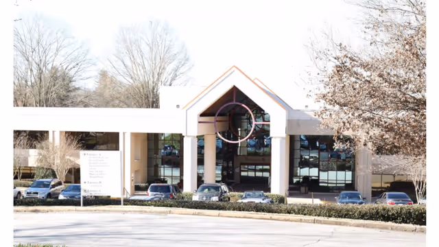 Front exterior view of a building with a peaked roof entrance supported by columns, large glass windows, several parked cars in front, and leafless trees around the parking area.