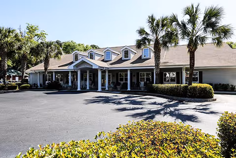 Front exterior view of a single-story building with a covered entrance supported by white columns, surrounded by palm trees and bushes under a clear blue sky.
