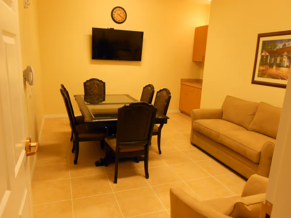 Small common room with a glass-top dining table and chairs, a wall-mounted TV and clock, and a beige sofa.