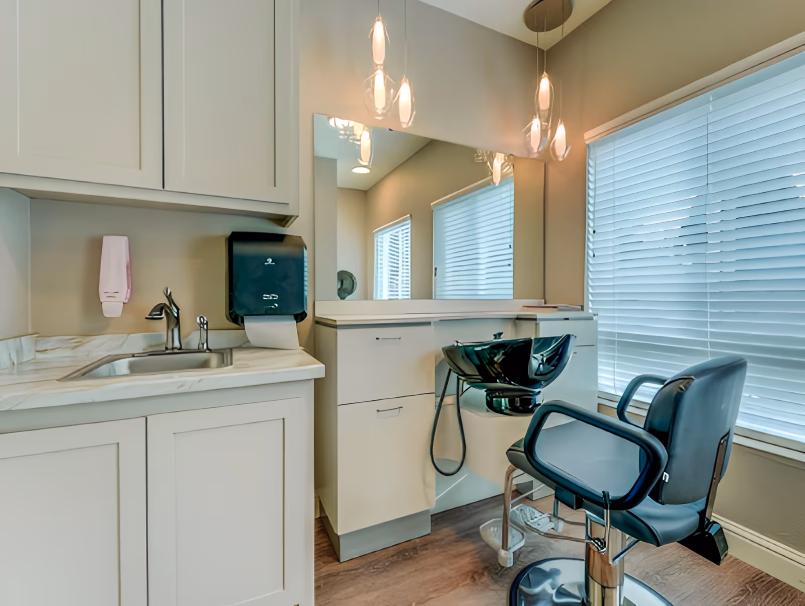 Interior view of a hair washing station in a salon area with a black shampoo bowl and a black salon chair. There is a large mirror on the wall, pendant lights hanging from the ceiling, a window with blinds, and white cabinetry with a sink and soap dispenser.