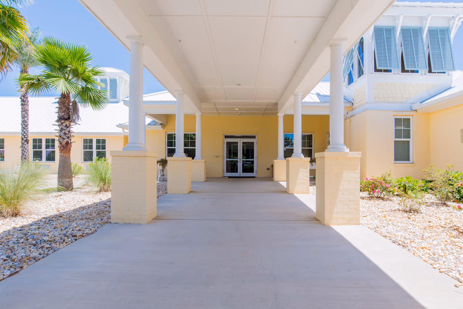 Covered entrance walkway to a light yellow building with white columns and a white roof, surrounded by landscaping with palm trees, bushes, and rocks under a clear blue sky.
