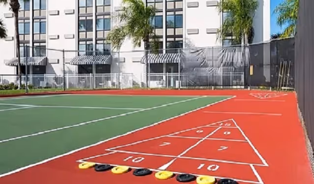 Outdoor shuffleboard court with numbered scoring zones and shuffleboard discs lined up at the starting area. In the background, there is a multi-story building with windows and striped awnings, along with some palm trees.