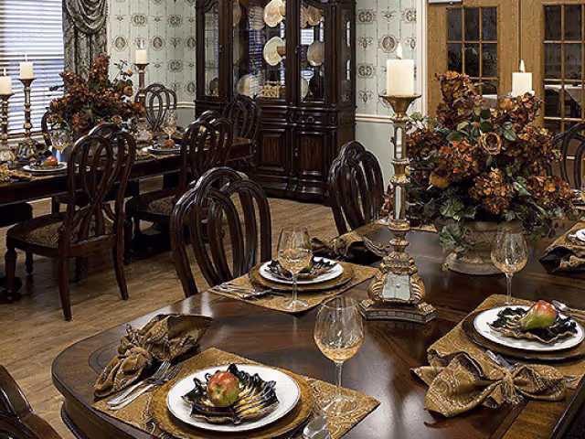 A formal dining room with dark wooden tables and chairs set with plates, wine glasses, and folded napkins. The tables have decorative centerpieces with flowers and tall candle holders. A large china cabinet with plates is visible against the wall, and the room has patterned wallpaper and wooden flooring.