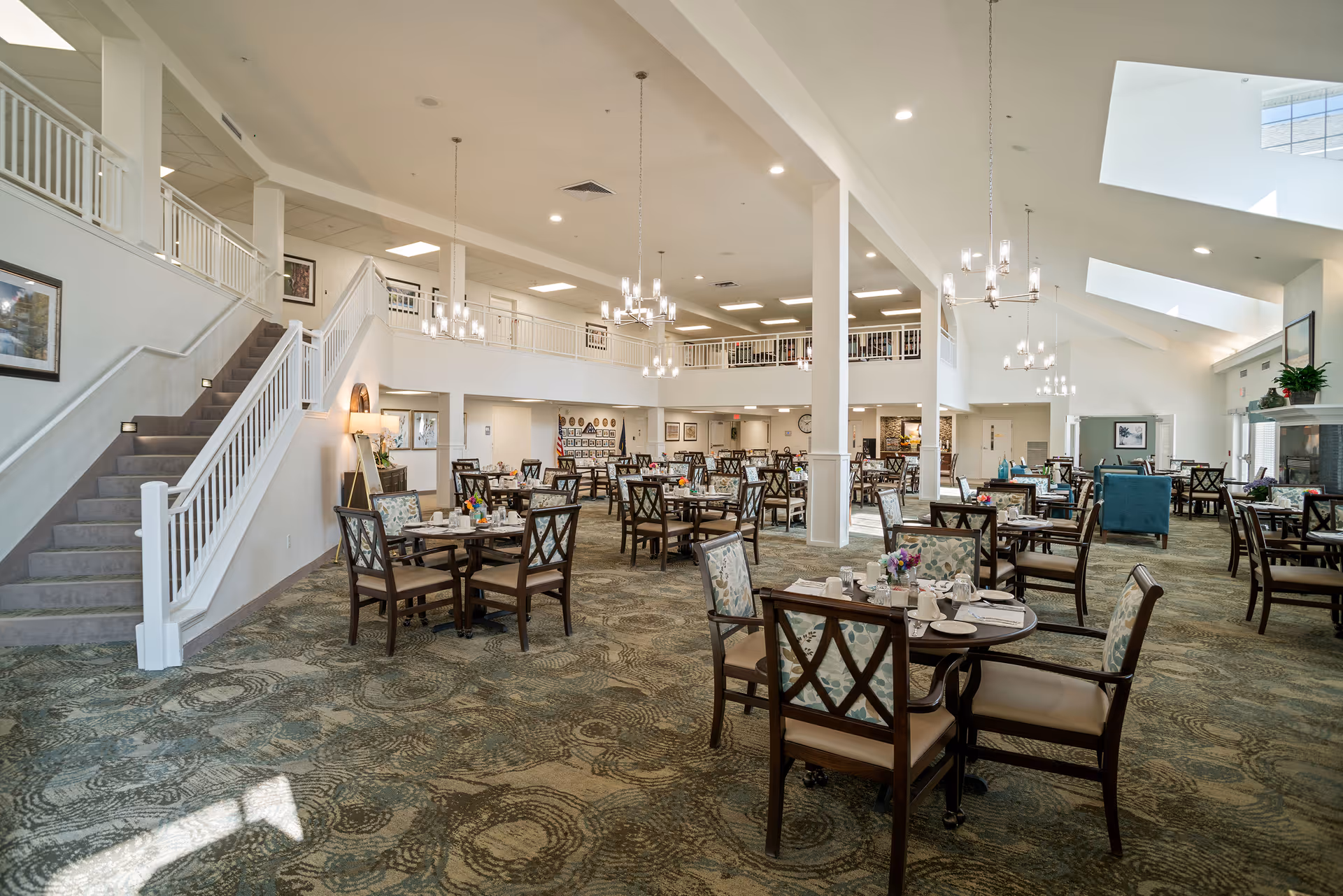 Spacious senior living dining room with multiple set tables, high ceilings, chandeliers, and a staircase leading to an upper balcony.