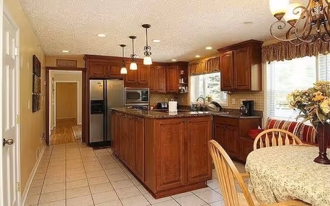 A spacious kitchen with wooden cabinets and a large central island with a granite countertop. The kitchen features stainless steel appliances including a refrigerator and microwave. There is a round dining table with a floral tablecloth and wooden chairs near a window with curtains. The floor is tiled, and the room is well-lit with ceiling lights and pendant lights above the island.