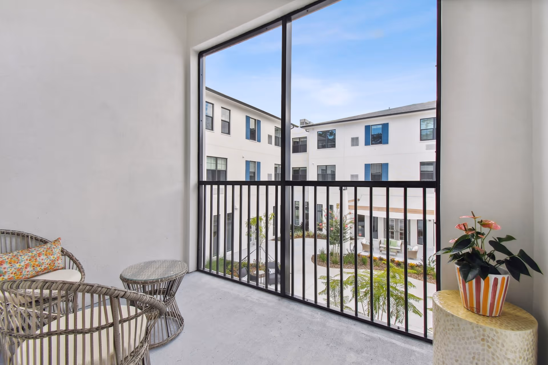 Screened balcony with wicker chairs and a potted plant overlooking the courtyard of a multi-story senior living building.