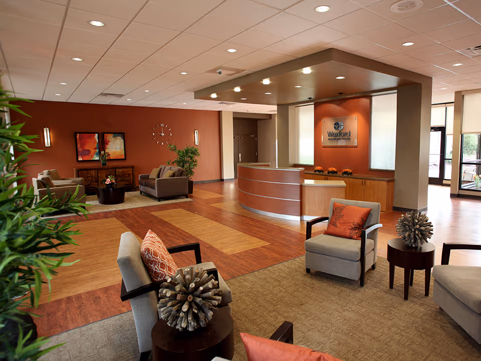 A spacious and well-lit reception area of Wexford Healthcare Center featuring a curved wooden reception desk, comfortable seating with armchairs and cushions, decorative plants, a wall clock, and framed artwork on a warm-toned wall.