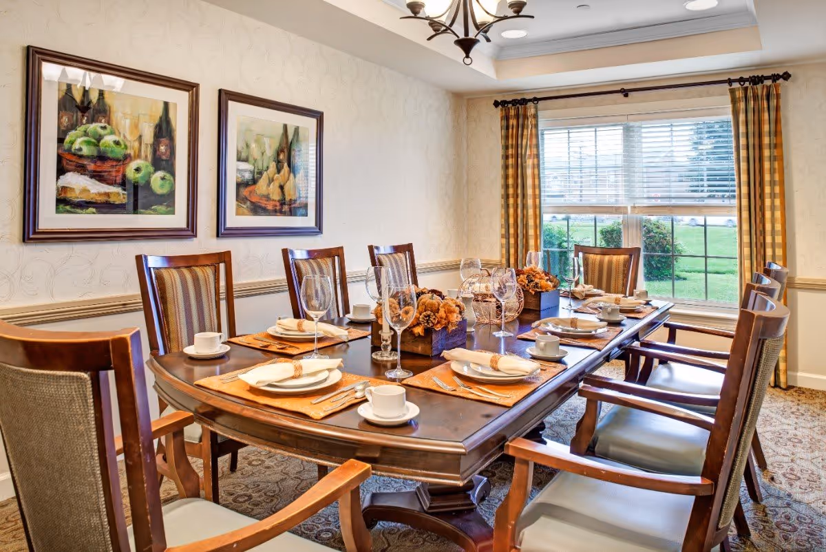A well-lit dining room with a large wooden table set for eight people. The table is arranged with white plates, cups, napkins, silverware, and wine glasses. There are decorative centerpieces with flowers and a candle holder. The room has patterned carpet, beige walls with two framed paintings of fruit, a window with yellow plaid curtains, and a chandelier overhead.
