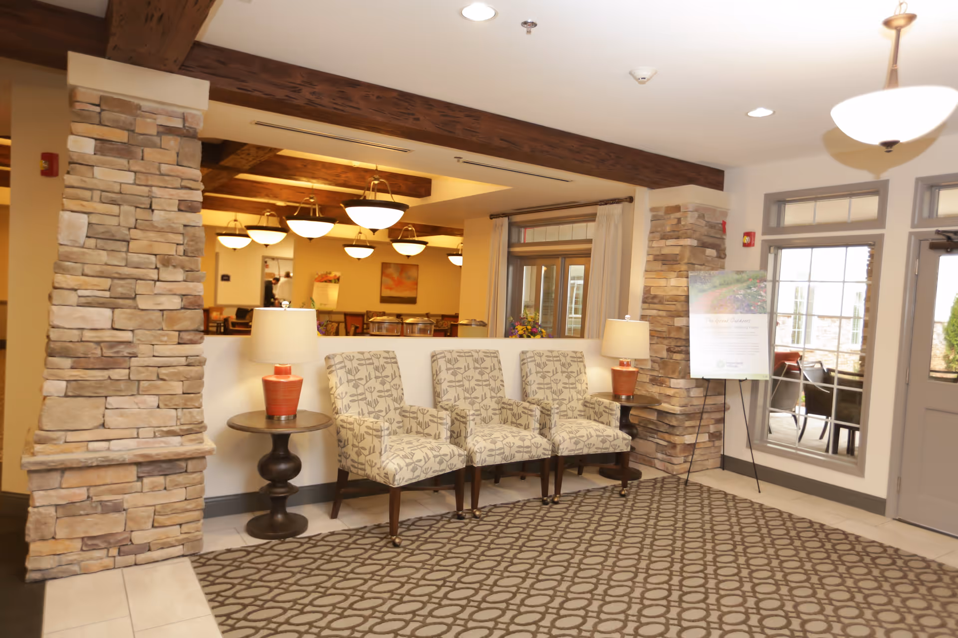 A bright interior reception area with three patterned chairs between stone columns, side tables with lamps, and pendant lights overlooking a dining area.