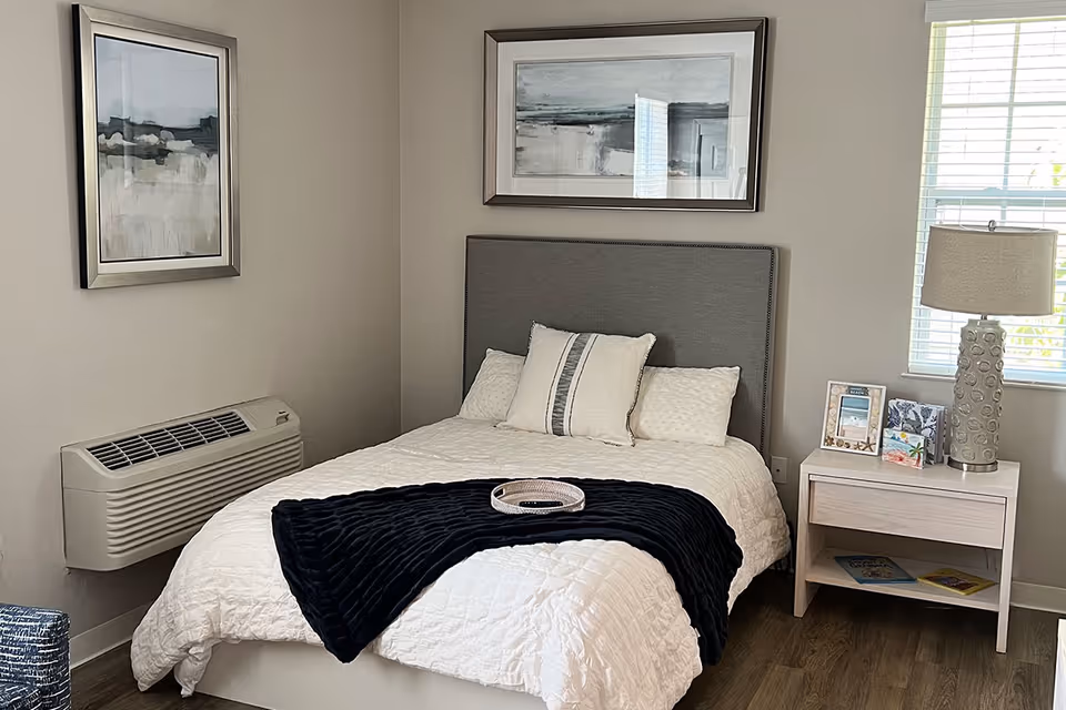 A neatly made bed with a gray upholstered headboard, white bedding, and a dark blue throw blanket folded at the foot. There are three pillows on the bed. On the right side, there is a light-colored nightstand with a decorative lamp, framed pictures, and some books underneath. Two abstract paintings hang on the walls, one above the bed and one on the adjacent wall. A window with blinds is partially visible on the right side, and a wall-mounted air conditioning unit is on the left wall. The room has wooden flooring and neutral-colored walls.