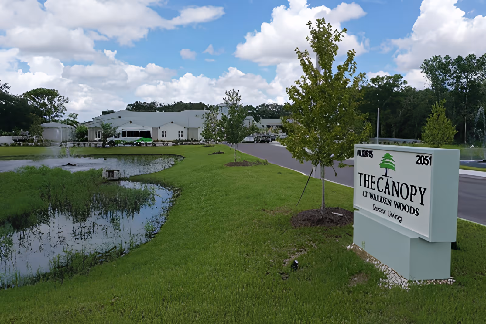 Sign for 'The Canopy at Walden Woods' on a grassy lawn by a pond with the facility building and trees in the background.