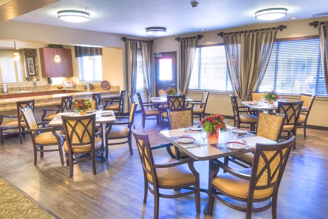 A bright and inviting dining room with several tables set for meals. Each table has four chairs with cushioned seats and backs, and is decorated with a floral centerpiece. The room features large windows with curtains, wooden flooring, and overhead lighting fixtures. A counter area with additional seating is visible in the background.
