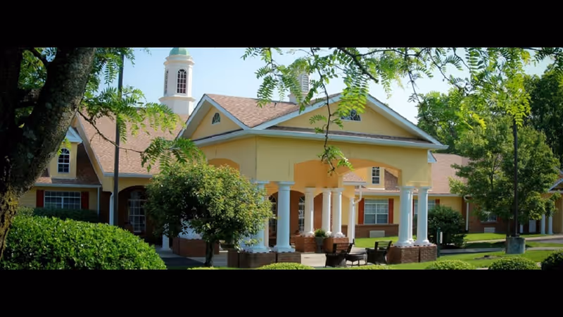 Exterior view of a senior living facility building with a yellow facade, white columns, and a small cupola on the roof. The building is surrounded by green trees and bushes, with outdoor seating visible near the entrance.