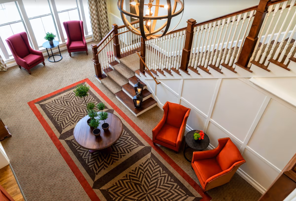 View from above of a cozy seating area in a senior living facility featuring a patterned carpet with red border, a round wooden table with potted plants, two red armchairs with a small black side table holding flowers, two purple armchairs near large windows with curtains, a staircase with wooden handrails and white balusters, and a large decorative hanging light fixture.