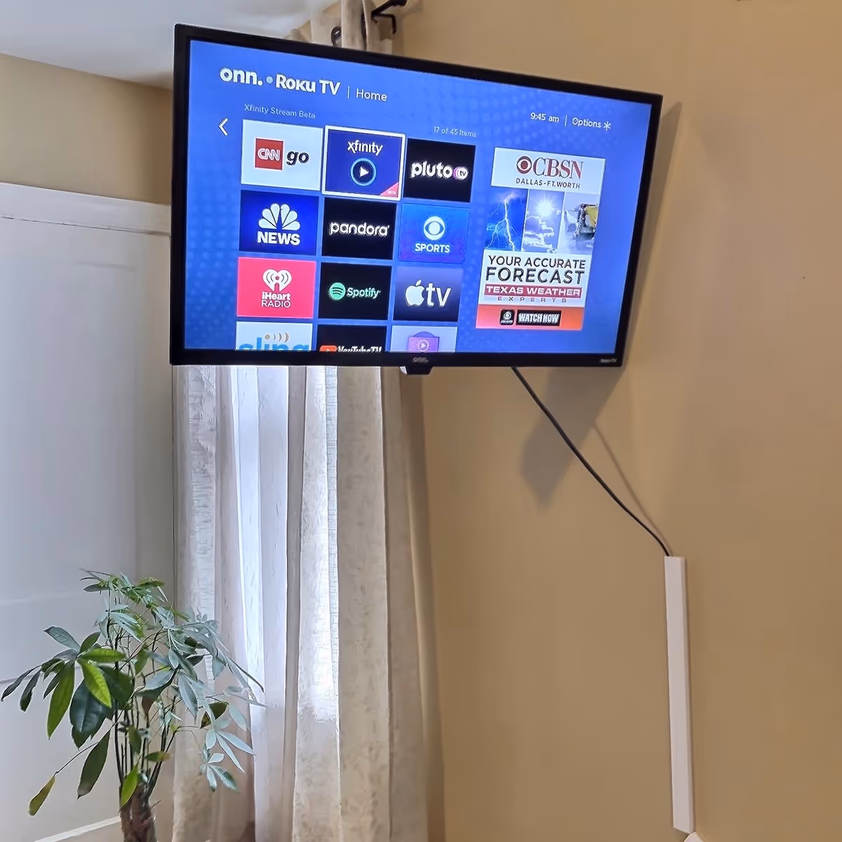 Wall-mounted TV showing a Roku home screen above a curtained window with a potted plant below.