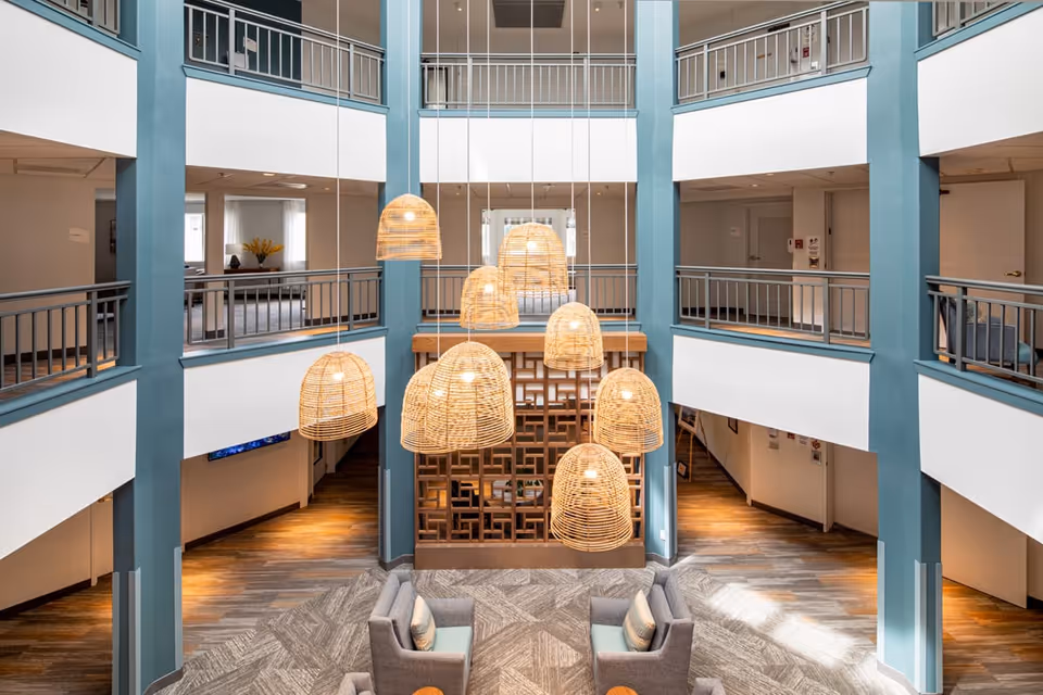 Interior view of a multi-level common area in a senior living facility with hanging wicker pendant lights, blue and white walls, railings on the upper floors, and seating areas with gray chairs and small tables on the ground floor.
