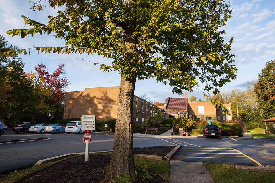 Exterior view of Clovernook Health Care LLC building with a parking lot in front, several parked cars, a large tree in the foreground, and a sign indicating nursing center entrance and ambulance parking. The scene is set on a clear day with some clouds and surrounding trees showing autumn colors.