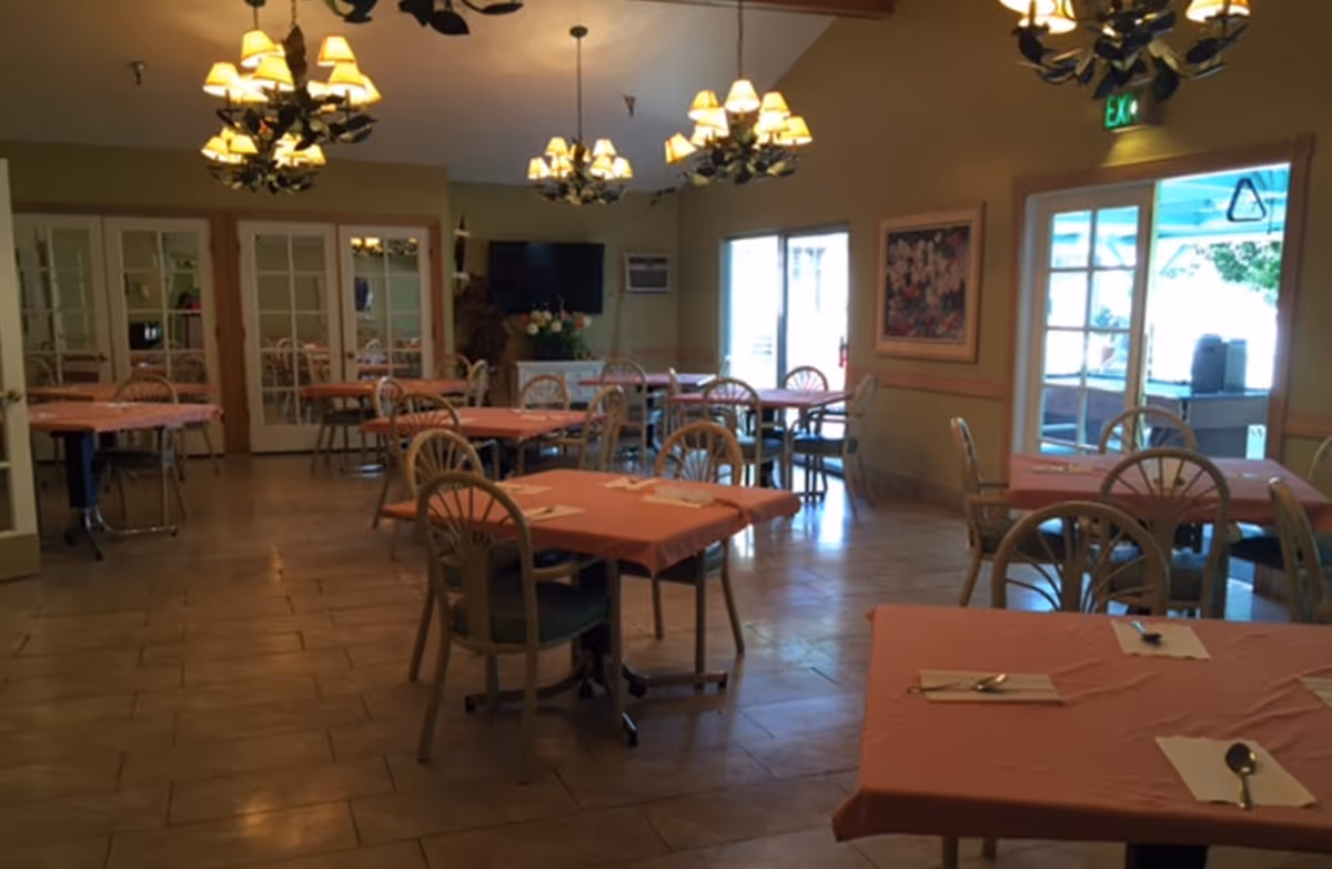A dining room with multiple tables covered in pink tablecloths, each set with napkins and silverware. The room features several chandeliers hanging from the ceiling, large windows letting in natural light, and a television mounted on the far wall. There are chairs around each table and a painting on one wall.