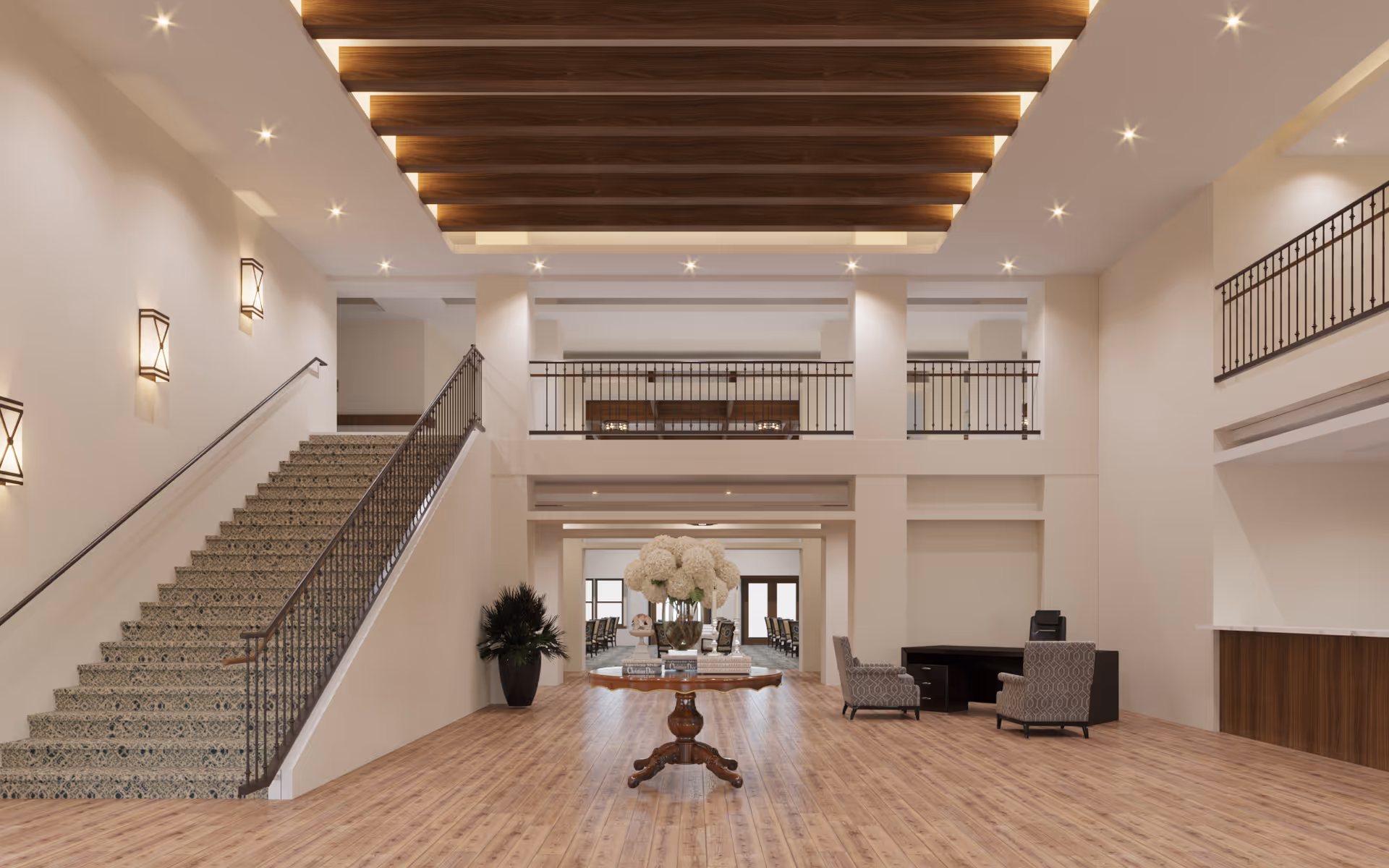 Spacious and well-lit senior living facility lobby with wooden flooring, a decorative wooden ceiling with recessed lighting, a staircase with patterned steps and metal railing on the left, a round wooden table with a large floral arrangement in the center, two upholstered chairs and a desk on the right, and a balcony railing on the upper level.