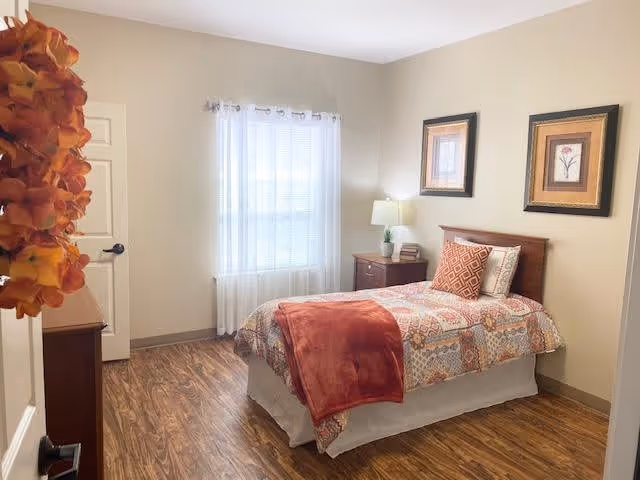 A cozy bedroom with a single bed covered in a patterned quilt and a red throw blanket. There is a wooden nightstand with a lamp and a small plant next to the bed. Two framed pictures hang on the beige wall above the bed. A window with sheer white curtains lets in natural light. The floor is wood, and a fall-themed wreath is partially visible on the door.