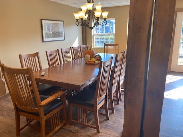 A dining room with a large wooden table surrounded by ten wooden chairs with black cushions. A chandelier with multiple lights hangs above the table. There is a decorative centerpiece on the table with pumpkins and gourds. The room has wooden flooring, a framed picture on the wall, and windows letting in natural light.