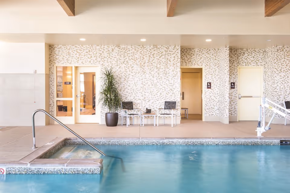 Indoor swimming pool with a handrail, seating area, potted plant, and doors along a tiled wall.