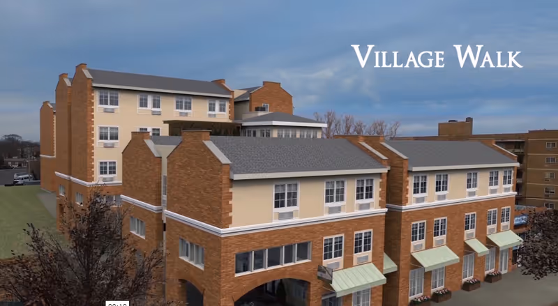 Multi-story brick and stucco senior living building with arched walkways and green awnings under a cloudy sky, with 'Village Walk' text in the corner.