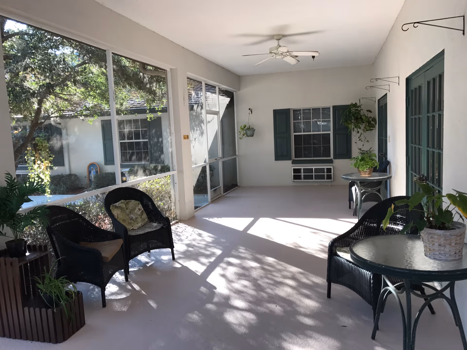 A screened-in porch area with black wicker chairs and glass-top tables, decorated with potted plants. The porch has large windows looking out to greenery and a ceiling fan overhead.
