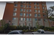 Multi-story brick residential building with rows of windows, shrubs along the sidewalk, and parked cars in front.