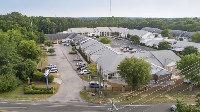 Aerial view of a single-story L-shaped senior living/post-acute rehabilitation complex with parking, landscaping, and a road with power lines in front.