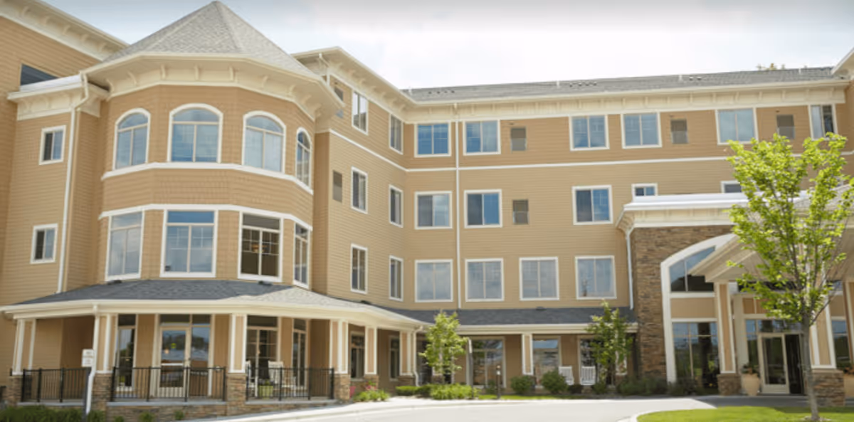 Exterior view of a multi-story senior living facility building with beige siding, large windows, and a covered entrance. There are small trees and shrubs planted around the building and a paved driveway in front.