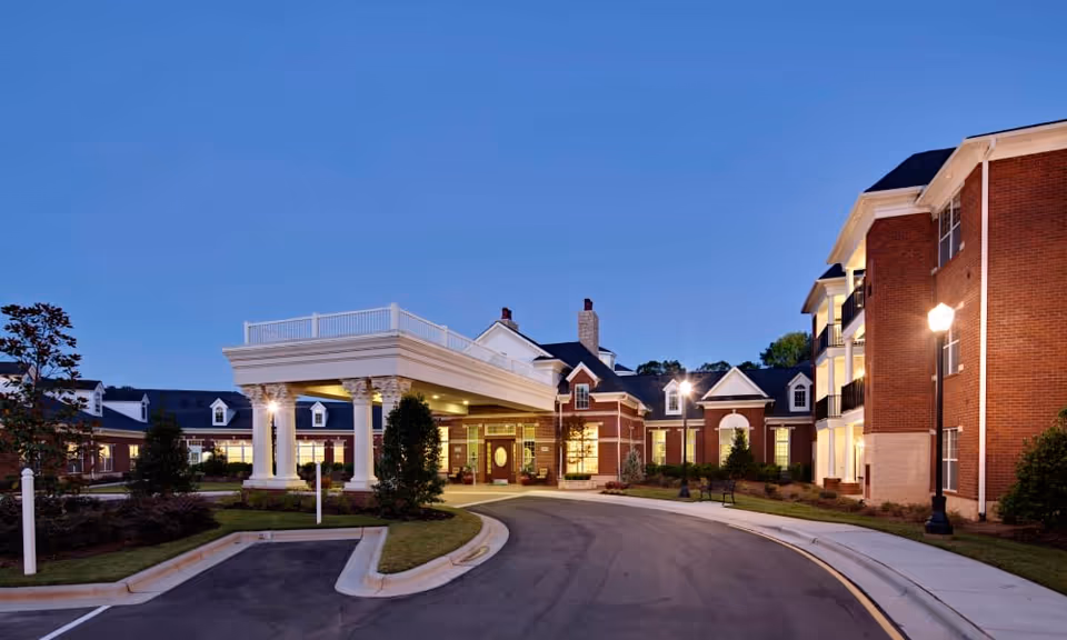 A brick senior living building's front entrance with a white-columned covered drive, curved driveway, and exterior lighting at twilight.
