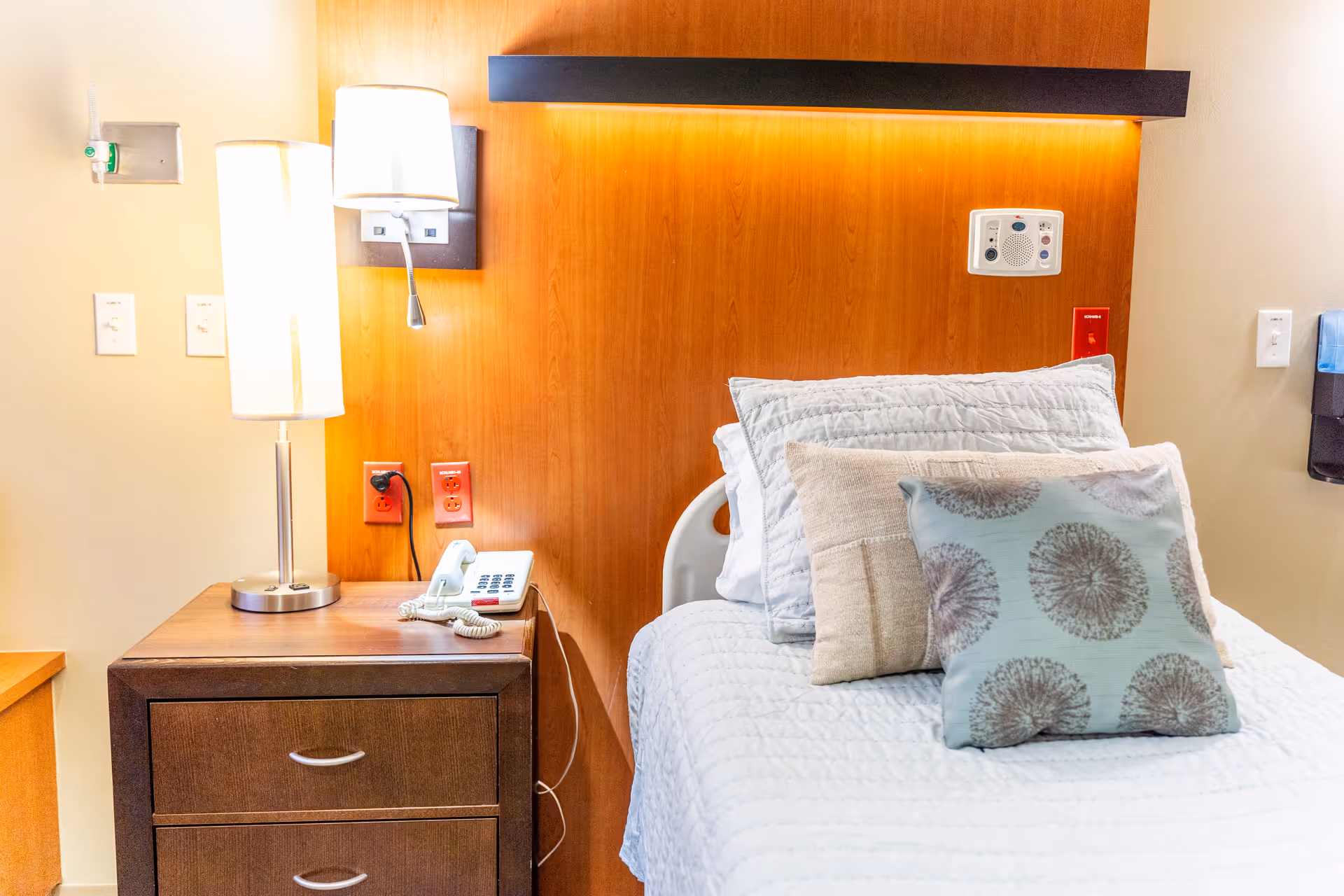 A neatly made hospital-style bed with white bedding and three decorative pillows in different patterns. Next to the bed is a wooden nightstand with two drawers, a modern lamp, and a white telephone. The wall behind the bed has a wooden panel with a mounted light fixture and medical equipment outlets.