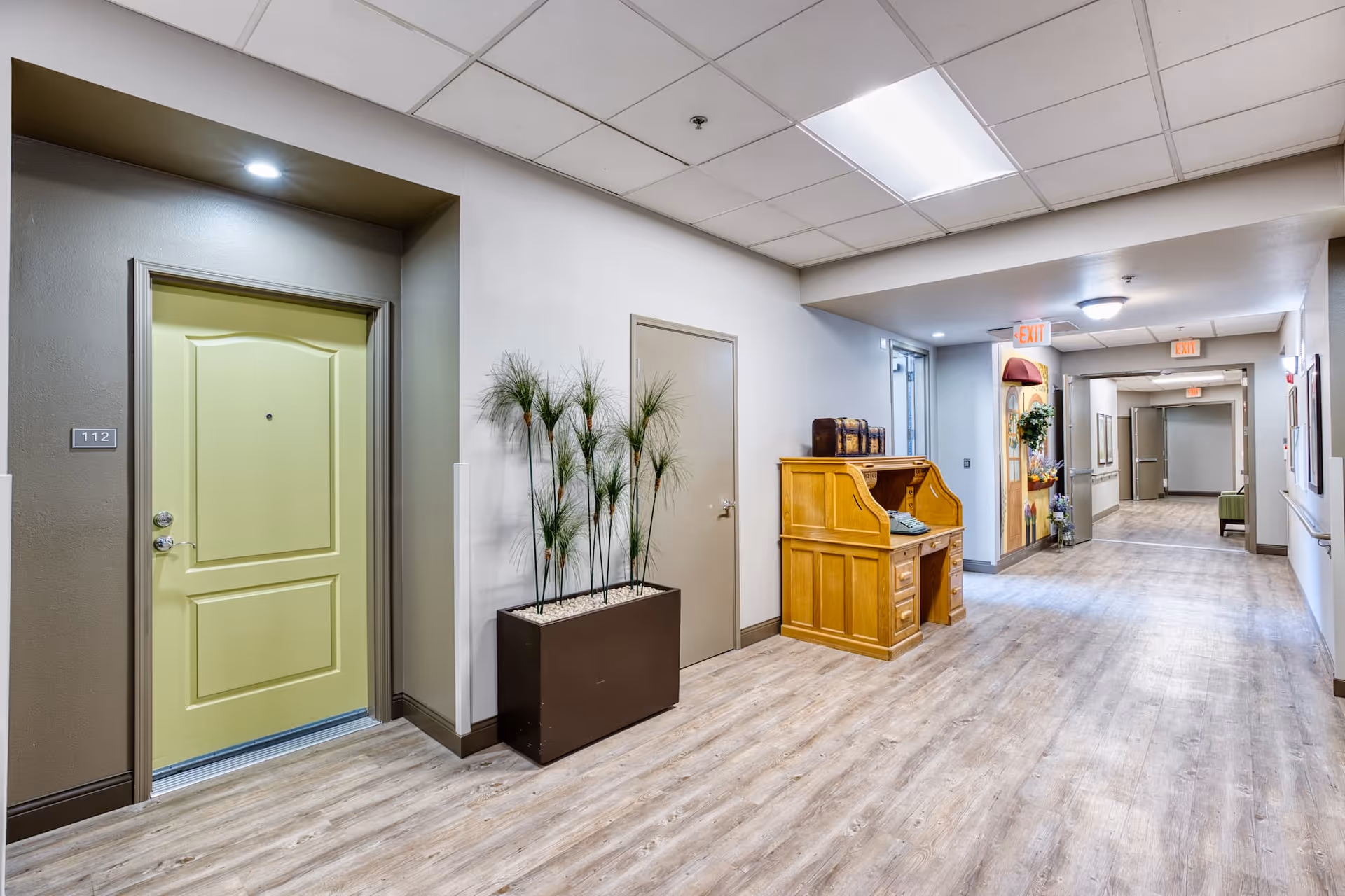 A clean, well-lit hallway in a senior living facility with light wood flooring and white walls. On the left side, there is a light green door labeled 112 next to a tall planter with green plants. Further down the hallway, there is a wooden desk with a vintage typewriter on top. The hallway extends into the distance with exit signs visible on the ceiling.
