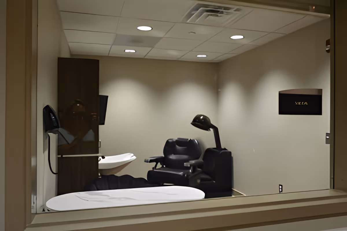 Interior view of a small salon or hair care room with a black salon chair, a hair dryer hood, a white sink, and a cabinet against beige walls and a white ceiling with recessed lighting.