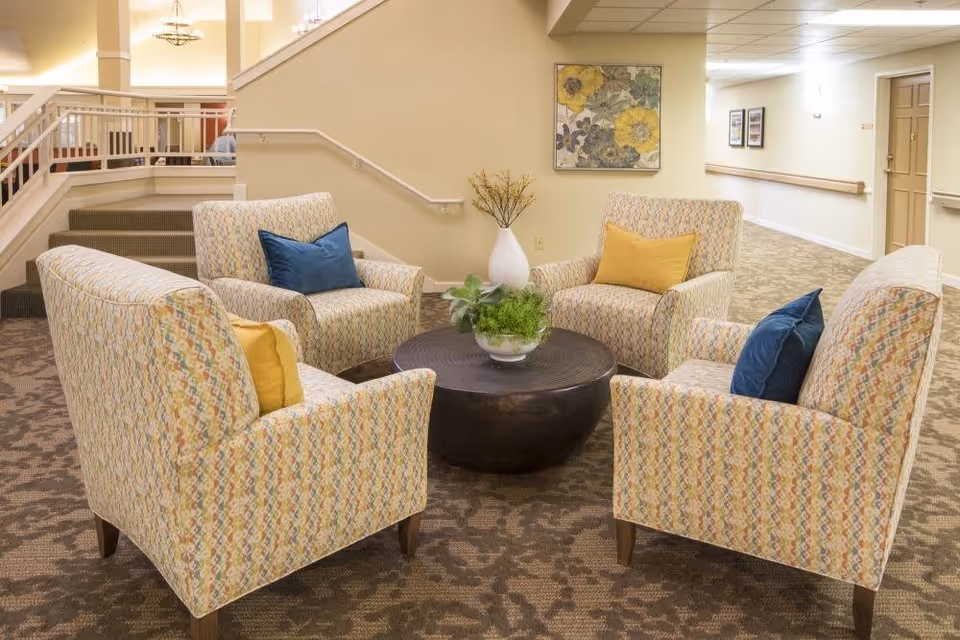 A cozy seating area in a senior living facility with four patterned armchairs arranged around a round dark wooden coffee table. The chairs have blue and yellow accent pillows. There is a vase with flowers on a small table against the wall, and framed artwork hanging above it. The area is carpeted and located near a staircase and hallway.