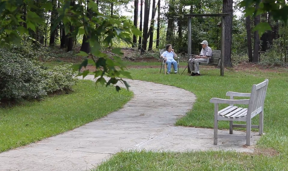 A winding paved pathway in a green outdoor area with trees and bushes. Two elderly people are sitting and talking; one is seated on a chair and the other on a wooden swing. There is also an empty wooden bench near the pathway.