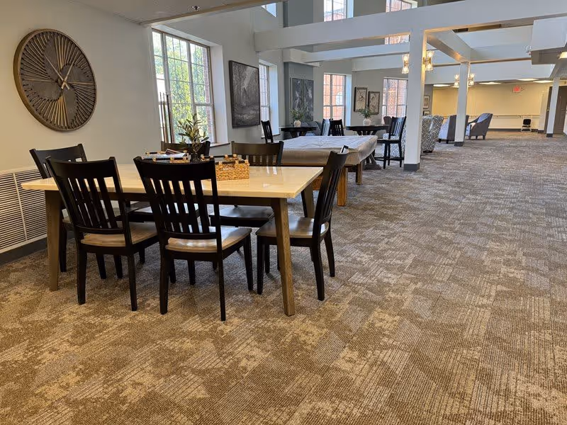 A spacious interior room with multiple tables and chairs arranged for seating. The room has large windows letting in natural light, carpeted floors, and neutral-colored walls. Decorative wall art and a large clock are visible on the walls. The space appears to be a common area or lounge in a senior living facility.