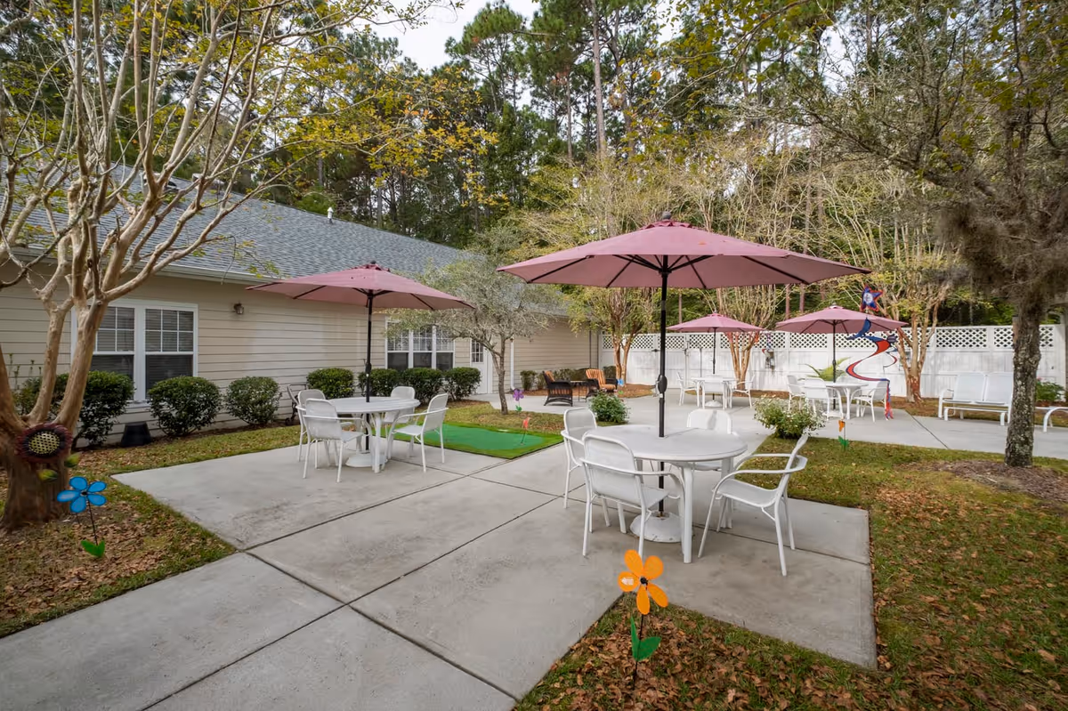 Outdoor patio area with several round white tables and white chairs, each table shaded by a pink umbrella. The patio is surrounded by trees and bushes, with a beige building in the background. Colorful flower-shaped decorations are placed in the grass near the patio.