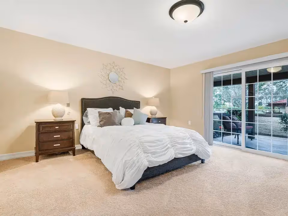 A bright bedroom with a large bed covered in white bedding and multiple pillows. There are two wooden nightstands on either side of the bed, each with a lamp. A decorative mirror hangs above the headboard. The room has beige walls and carpeted floor. Sliding glass doors lead to an outdoor patio with chairs and a table visible outside.