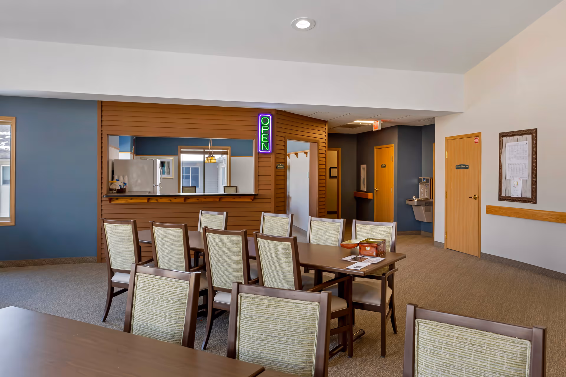 Interior view of a senior living facility dining area with multiple tables and chairs. There is a serving window with a neon green and purple 'OPEN' sign above it. The walls are painted in blue and white, and there are doors and a water fountain visible in the background.
