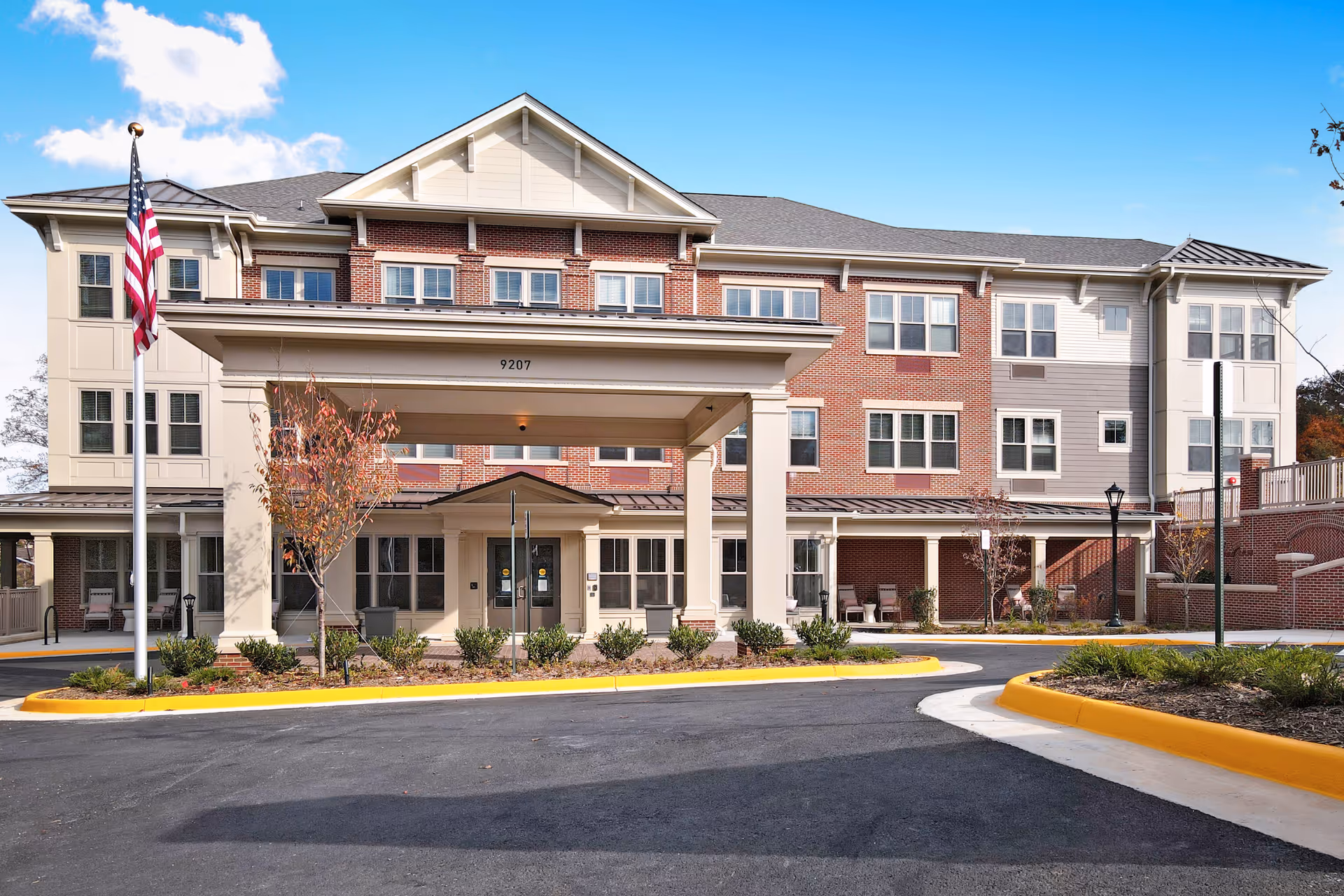Front exterior view of a three-story senior living facility building with a covered entrance, an American flag on a flagpole, landscaped bushes and trees, and a clear blue sky with some clouds.