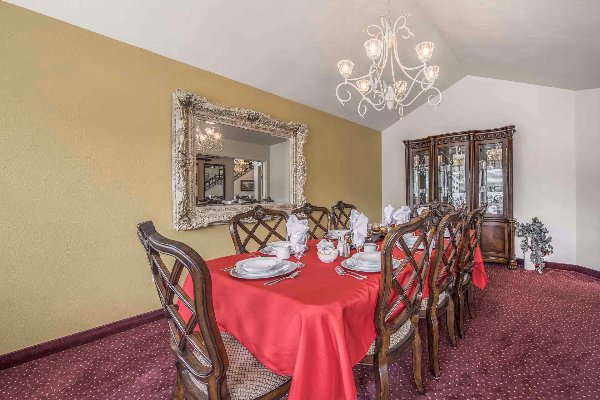 Formal dining room with a long table set with white dishes and a red tablecloth, a chandelier overhead, an ornate wall mirror, and a china cabinet.