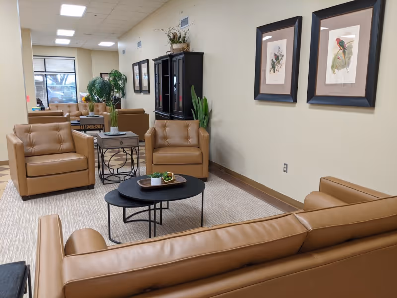 A cozy living room area in a nursing home with tan leather armchairs and a sofa arranged around two black nested coffee tables. The tables have decorative items including a small plant and a tray. The room has beige walls adorned with framed bird artwork, a black cabinet with plants on top, and a large window letting in natural light. The floor is covered with a light-colored rug and the ceiling has recessed lighting.