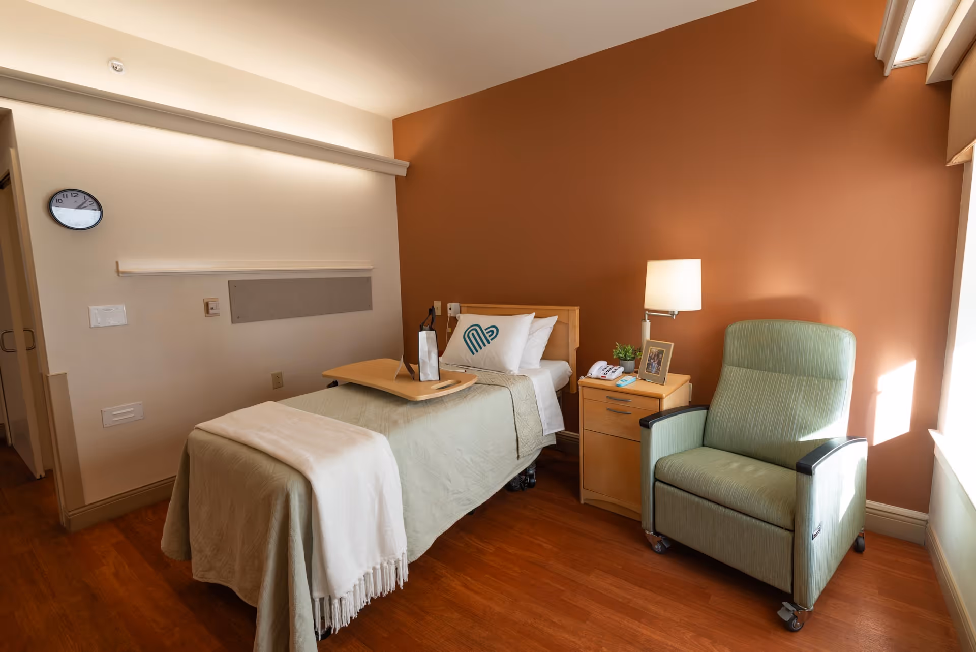 A tidy single-occupancy senior care room with a bed, bedside table and lamp, and a recliner chair against an orange accent wall.